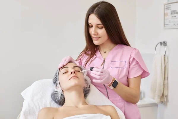 Attractive female laying with closed eyes on cosmetic chair in beauty parlor, having medical cap on head and white towel. Charming adorable cosmetologist in pink lab coat applying moisturizing oil.