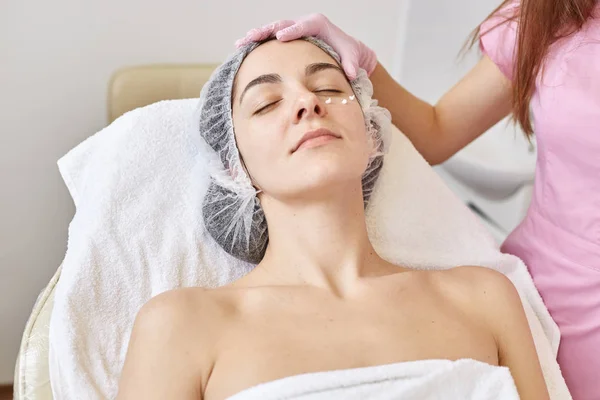 Indoor shot of satisfied relaxed woman closing her eyes while laying on coach, getting cosmetic service, having nourishing cream under eyes, spending time in spa salon. People and cosmetology concept.
