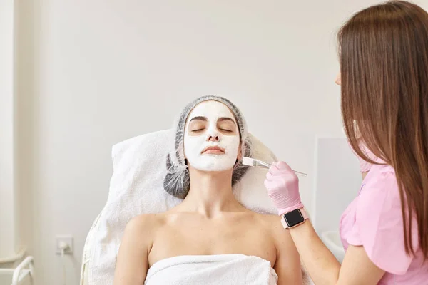 Indoor shot in beauty parlor of client and beautician with professional brush applied spa cream on woman face. Peaceful calm woman laying on coach with closed eyes and white mask on face. Spa concept.