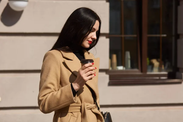 Joven hermosa mujer en abrigo beige con bolso de mano caminando por la calle isdolated sobre el fondo de la cafetería, tiene el pelo largo y oscuro, tiene manicura de color rojo brillante, señora de negocios después de la reunión. Concepto empresarial . — Foto de Stock