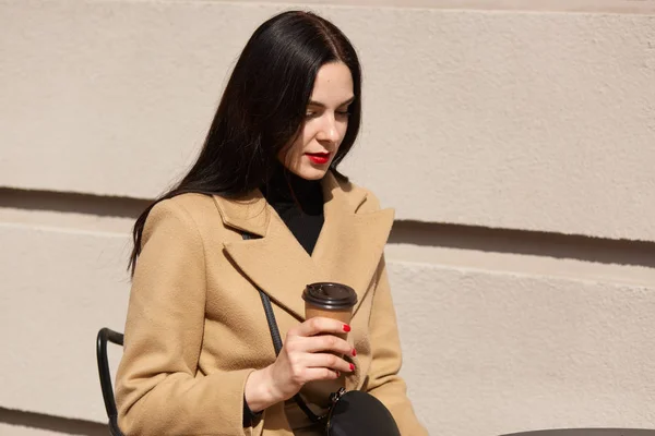 Encantadora joven morena con pelo largo y oscuro usando abrigo, sentada en la mesa en la cafetería de la calle y bebiendo café o té, atractiva mujer tiene una expresión facial reflexiva, preocupada por su trabajo . — Foto de Stock