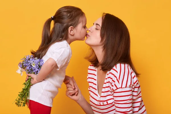 Young beautiful lady in striped shirt holds her daughter's hand and kisses her. Little child with bouquet of flowers behinds back congratulates her mom with mother's day. Family and holiday concept. — Stock Photo, Image
