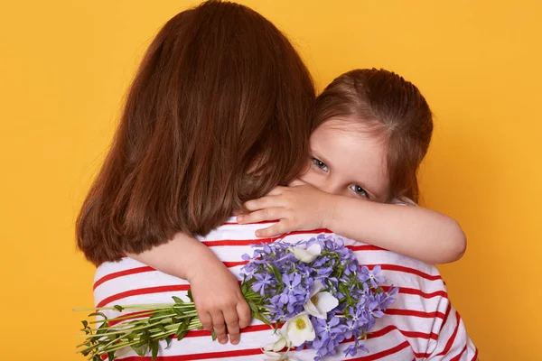 Beautiful young woman and her charming little daughter hugging indoor. Child congratulates mom with Mother's Day, gives flowers, mummy stands backwards wearing striped shirt. Holiday concept. — Stock Photo, Image