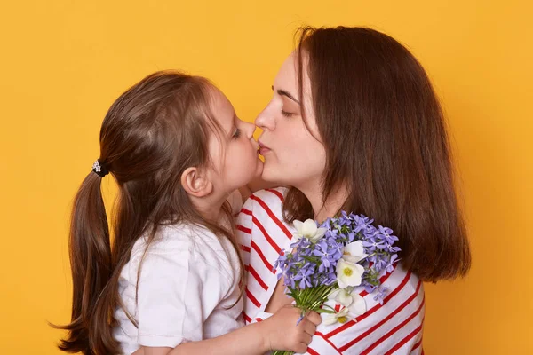 Studio shot of mother and daughter kissing and hugging, little girl standing with bouquet of blue florets for mommy. Dark haired woman poses with her child. Happy Mother's Day! Holiday concept. — Stock Photo, Image
