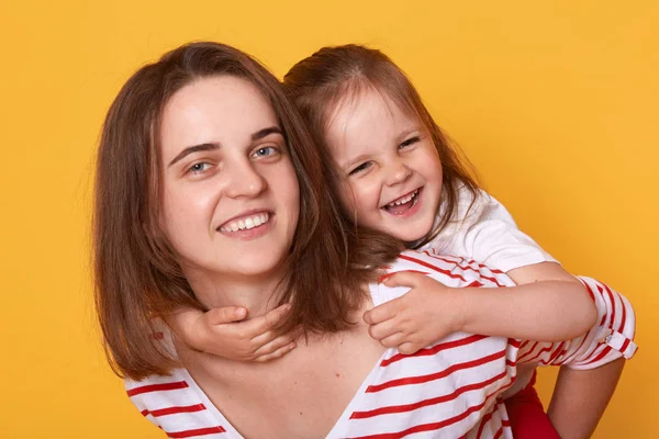 Daughter hugging mother from back, affectionate woman with toothy smile holds happy toddler, kid embracing mommy, family celebrating Mother's Day, posing isolated on yellow studio background. — Stock Photo, Image