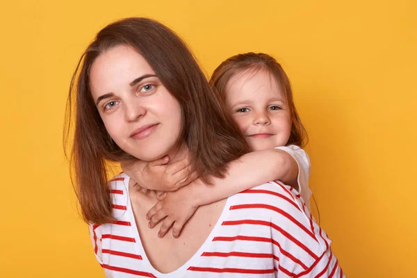 Happy mother's day! Child daughter hugging mom from behind. Mum wearing striped shirt and little girl looking at camera with happy facial expressions. Family, holiday and togetherness concept. — Stock Photo, Image