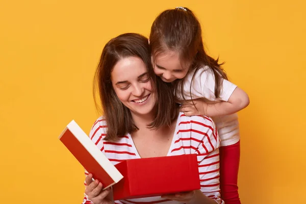 Family, child, holiday and Mother's Day concept. Smiling young woman in striped shirt sitting with her toddler daughter and opening red box, present for holiday, liitle girl in t shirt and trousers.