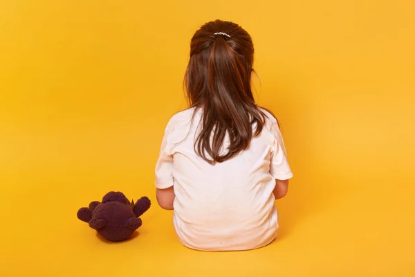 Little girl sitting backwards with brown teddy bear, toddler wearing white t shirt offended by someone, being in bad mood, feels sad and upset, posing isolated over yellow background. Emotions concept — Stock Photo, Image