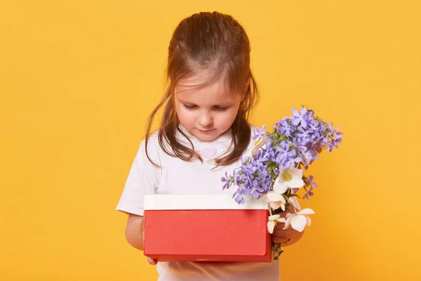Dolce bambina che tiene la scatola rossa con regalo e mazzo di fiori, il bambino si preoccupa mentre si prepara a congratularsi con la sua mamma con la festa della mamma, guardando il suo regalo. Concetto bambini e vacanze . — Foto Stock