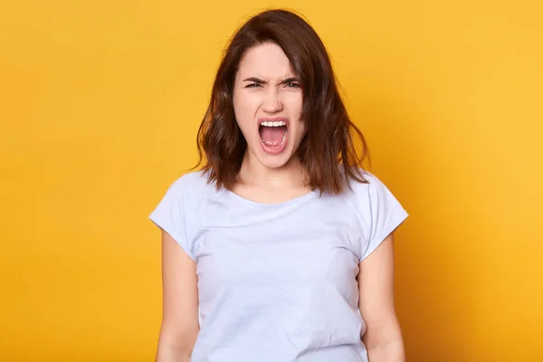 Gritando mujer enojada emocional aislado sobre fondo de estudio amarillo. Retrato de media longitud de una joven morena vestida con una camiseta blanca casual. Emociones humanas y concepto de expresión facial . — Foto de Stock