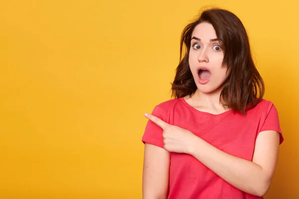 Studio shot de mujer negra asombrada señala a un lado con el pulgar, ha emocionado la expresión, sorprendido al escuchar nueva pista, lleva camiseta negra casual, aislado sobre la pared del estudio amarillo. Cielos, ¿qué noticias — Foto de Stock