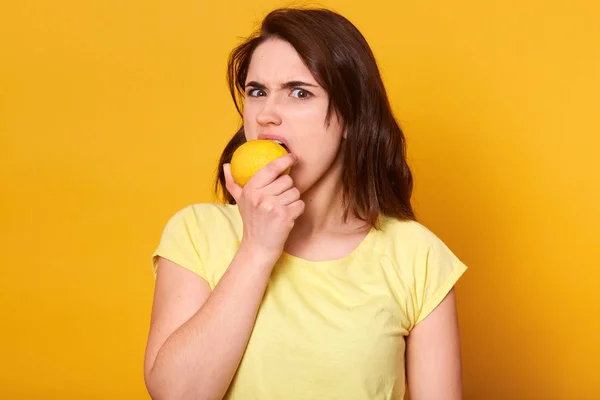Mujer joven disgustado en camiseta casual morder limón aislado sobre fondo amarillo en el estudio, necesita más vitaminas, mirando a la cámara con expresión facial descontenta, quiere algo sweter . — Foto de Stock