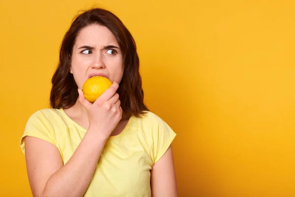 Vitaminas naturales y concepto de comida saludable. Indoor shot of young brunette woman bits lemon with discontented facial expession, looking airily aside, copy space for your promotion text or advertisment . — Foto de Stock