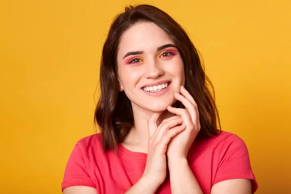 Indoor shot di bella femmina con pelle naturale perfetta e trucco luminoso nei colori rosa e arancio, guardando sorridente fotocamera, tiene le mani insieme vicino al viso, vestito t shirt rossa. Concetto bellezza . — Foto Stock