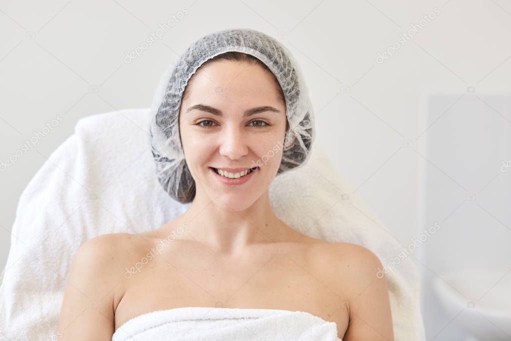 Close up portrait of charming good looking female posing over white wall in beauty parlor wearing medical cap and white towel, having positive facial expression, looks satisfied. Cosmetology concept.