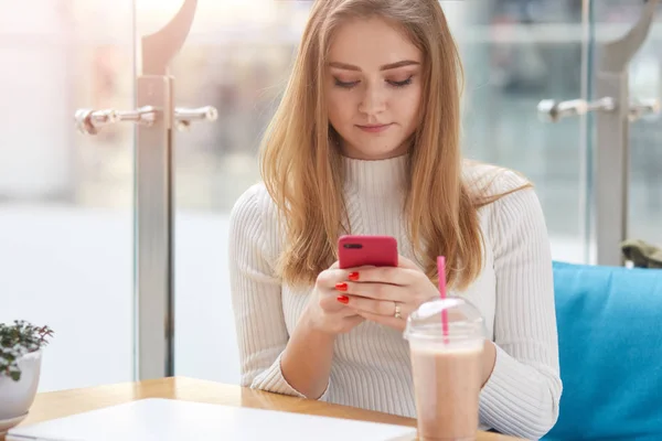 Mavi kanepede kafede oturan beyaz gömlekli çekici sarışın kadın, cep telefonu tutan, wi-fi kullanarak arkadaşları ile sohbet, kokteyl içme, birlikte vakit geçirmek için erkek arkadaşı bekliyor. — Stok fotoğraf