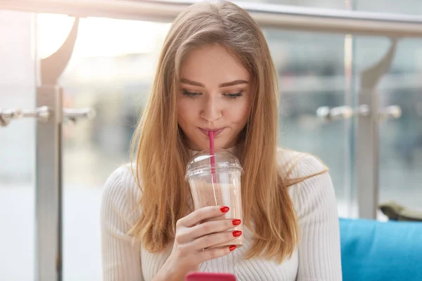 Close up retrato linda menina em camisa casual branca, tem cabelo loiro longe, beber coquetel sentado no café de verão. Foto interna de jovem estudante, olhando para baixo em seu telefone inteligente, lê algo . — Fotografia de Stock