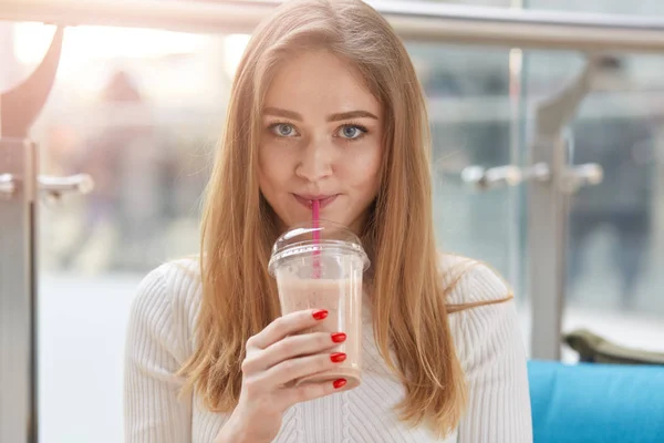 Plan intérieur d'une belle jeune femme buvant un cocktail au lait, regardant directement la caméra, tenant une tasse avec une moyenne fraîche à la main, appréciant le goût sucré de boire, portant une chemise décontractée blanche . — Photo