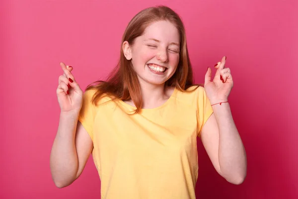 Allegro giovane bella ragazza con le dita incrociate, esprime il desiderio, sorride in generale, indossa casual t shirt gialla, modelli sopra parete rosa con spazio copia per le vostre informazioni. Donna speranze piano funzionerà . — Foto Stock