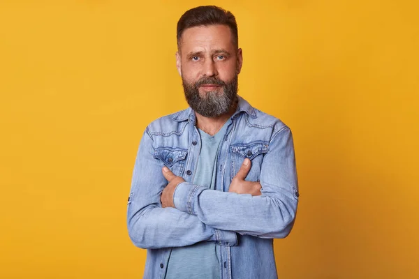Close up portrait of handsome middle aged Caucasian man with beard, posing isolated on yellow background, wears denim jacket and gray t shirt, has serious facial expression, keeps arms crossed. — Stock Photo, Image