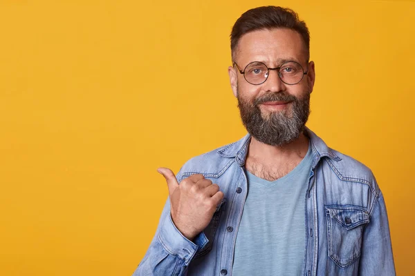 Happy optimistic handsome middle aged male with beard pointing aside with thumb and looking at camera, smiling man in denim jacket and gray t shirt. Copy space for promotin text or advertisment. — Stock Photo, Image
