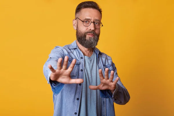Image of attractive middle aged man showing refusal gesture, has serious facial expression, wearing stylish denim jacket, gray t shirt and glasses, poses against yellow concrete studio background. — Stock Photo, Image
