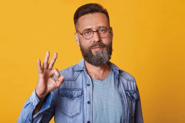 Studio shot of handsome man with beard wearing denim jacket, gray t shirt and spectacles, has positive facial expression, doing ok sign with hand and fingers, isolated over yellow studio background. — Stock Photo, Image