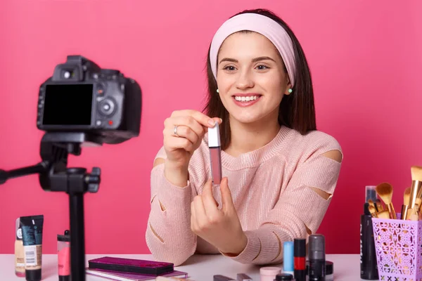 Sonriendo hermosa vlogger mostrando su nuevo lápiz labial a los suscriptores, se ve feliz, sosteniéndolo en ambas manos, posando aislado sobre fondo rosa en el estudio, demostrando nuevos productos cosméticos . — Foto de Stock