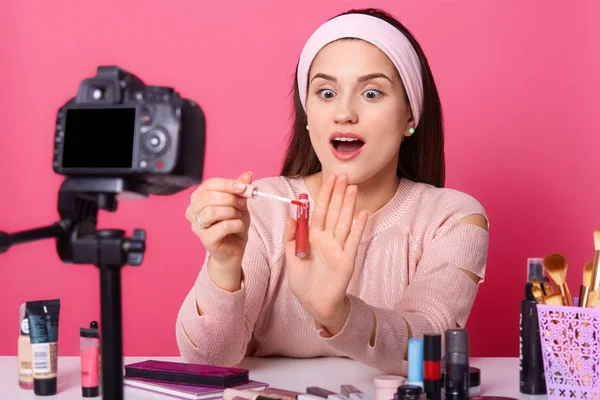 Imagen del joven bloguero sorprendido, lleva camisa casual y banda de pelo, hermosa mujer se sienta delante de la cámara digital, sosteniendo brillo de labios, mirándolo, impresionado por la calidad. Concepto de belleza . — Foto de Stock