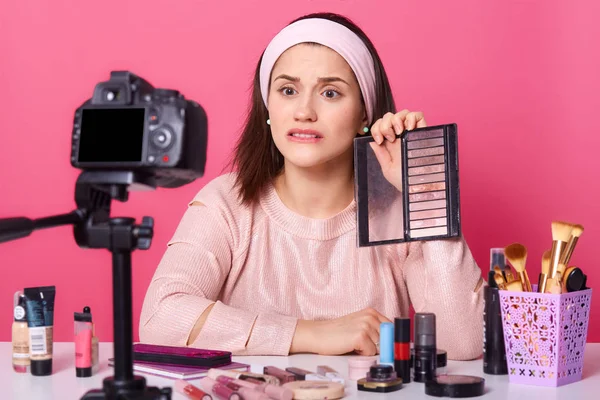 Indoor shot de blogger beaty atractivo decepcionado, sosteniendo paleta de sombra de ojos, demostración de la misma a los espectadores, posando aislado sobre fondo rosa en el estudio delante de la cámara, se ve molesto . — Foto de Stock