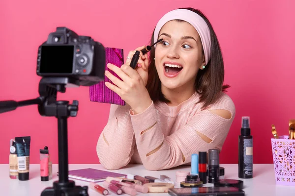 Mujer alegre con la boca abierta y los ojos haciendo tutorial para sus suscriptores, sosteniendo espejo rosa, aplicando rímel, haciendo maquillaje diario, estar de buen humor, grabación de vídeo en su cámara . — Foto de Stock