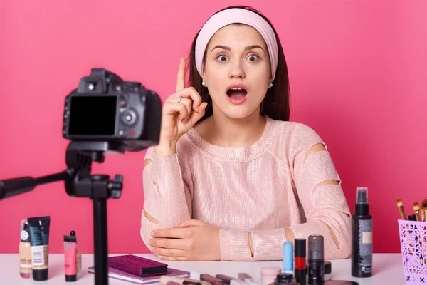 Sorpresa carina giovane donna creativa seduta a tavola piena di cosmetici, avendo fotocamera davanti al viso, alzando il dito, aprendo la bocca e gli occhi ampiamente, indossando oggetti casual di vestiti . — Foto Stock