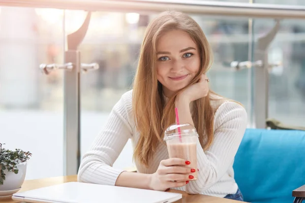 Sevimli sevimli femal görüntü doğrudan kameraya bakarak, kafeteryada öğle yemeği, milkshake plastik fincan tutan, masaya dizüstü koyarak, mutlu ve olumlu görünüyor, rahat kazak giyiyor. — Stok fotoğraf