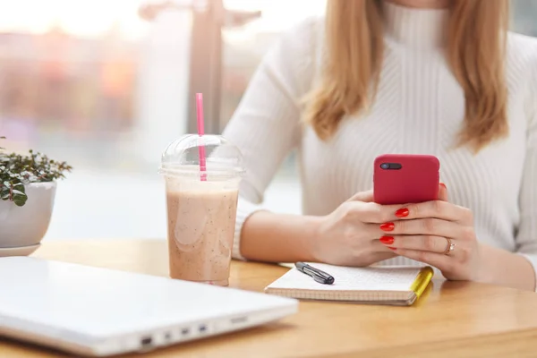 Felismerhetetlen nő a piros okos telefon a kezében, írja üzenetek, dolgozó online cafeteria, miután notebook, toll, műanyag csésze koktél és a fehér laptop az asztalra. Emberek és munkakoncepció. — Stock Fotó