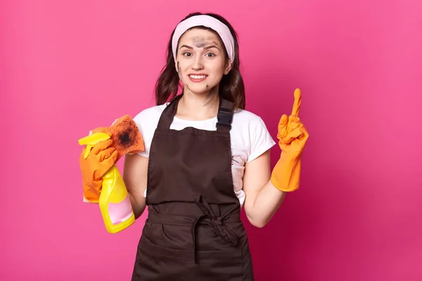 Retrato de enérgico limpiador sonriente lindo de pie con paño sucio y detergente, con guantes de color naranja, camiseta blanca, diadema y delantal marrón, levantando un dedo hacia arriba, con la cara sucia . —  Fotos de Stock