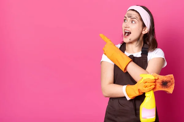 Energetic slender housewife with wide opened mouth, looking aside, raising hand, showing direction with forefinger, holding washcloth and detergent in one hand. Copy space for advertisement. — Stock Photo, Image