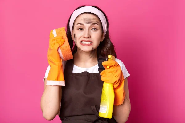 Limpiador emocional agotado de pie aislado sobre fondo rosa en el estudio, sujetando el paño cerca de la oreja y el detergente, con expresión facial desagradable, mirando directamente a la cámara con la cara sucia . — Foto de Stock