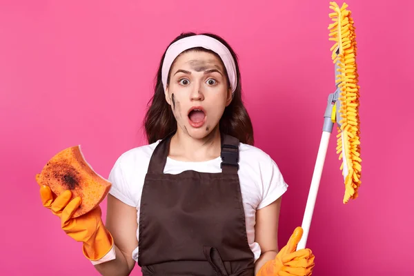 Portrait of shocked exhausted cute woman with wide opened mouth and eyes, feeling of amazement, holding broom and dirty washcloth, being impressed, standing with dirty face. Household concept. — Stock Photo, Image