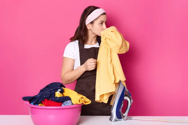 Young woman works as maid, wears t shirt, brown apron and hair band, standing near pink basin with clean linen isolated on rose background in photo studio, smells fresh clothes, enjoing pleasant odor.