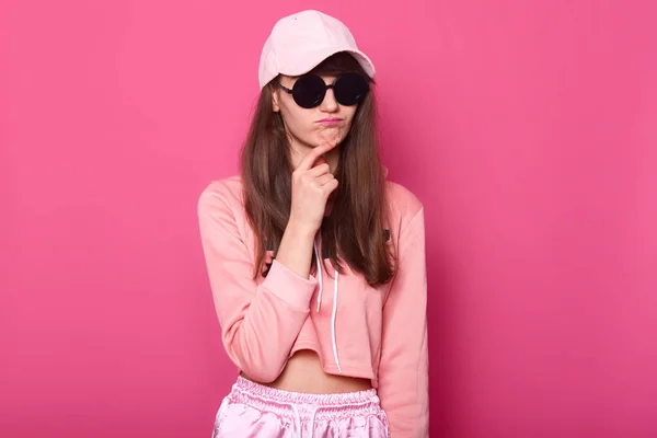 Srusio shot van stijlvolle tiener meisje dragen modieuze verkorte roze hoodie, GLB en zwarte subglasses, raakt haar kin met duivellijke facialexpression. Portret van de vrouw. People concept. — Stockfoto