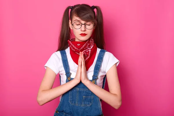 Close-up portret van kalm mooie Kaukasische vrouwelijke tiener met ponytails, houdt de ogen gesloten, bidt terwijl staande tegen roze muur, draagt wit t shirt, overalls, rode Bandana op nek en glazen. — Stockfoto