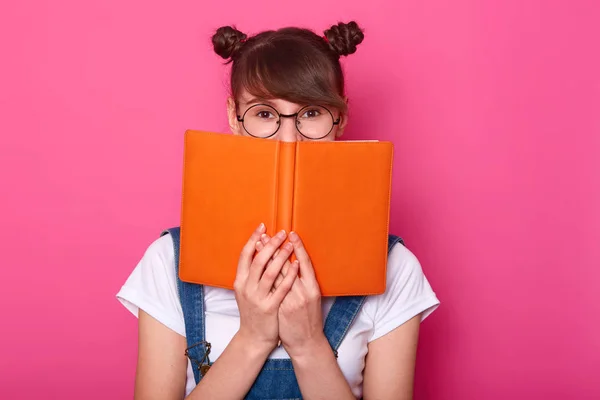 Chica pensativa feliz de pie aislado sobre fondo rosa en el estudio, sosteniendo cuaderno naranja, cubriendo la mitad de la cara, mirando atentamente a la cámara, teniendo racimos, usando ropa casual de moda . —  Fotos de Stock
