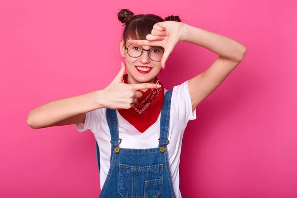 Estúdio interno tiro de fêmea energética alegre mostrando sinal de fazer fotos, colocando as mãos na frente de si mesma, sorrindo sinceramente, olhando diretamente para a câmera, poding isolado sobre fundo rosa . — Fotografia de Stock