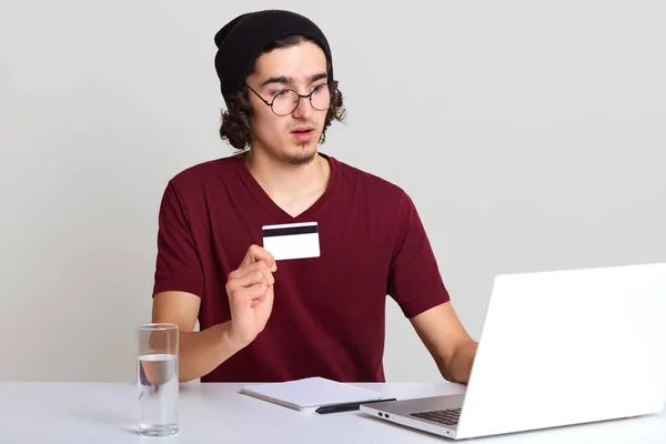 年轻帅哥穿着休闲的栗色T恤和黑色帽子，坐在打开的笔记本电脑前，显示信用卡，隔离在白色墙壁上，喝水，网上买东西，使用wirelwss互联网. — 图库照片