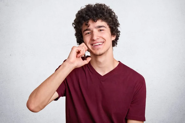 Close-up portret van jonge man met krullend haar, in kastanjebruine t shirt praten op de telefoon terwijl het kijken naar de camera geïsoleerd over witte studio achtergrond, heeft een aangenaam gesprek met zijn vriendin. — Stockfoto
