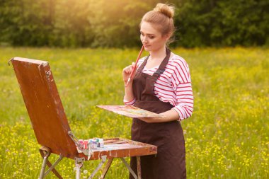 Genç sarışın kadın sanatçı, çizim ve düşünme, ilham arayan, açık havada poz, doğa, bayan soyulmuş gömlek ve kahverengi önlük giyen yakın portre kadar. Sanat ve yaratılış kavramı.