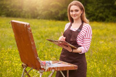 Sanatçı, açık havada resim çizer, elinde yağlı boya fırçası ve renk paleti tutarak yaratıcılığını hayata geçirdi. Büyüleyici gülümseme ile Bayan kameraya bakar, rahat giyen, manzara çizer.