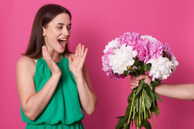 Sürpriz, mutluluk ile buket bakarak etkilendim genç kadın, şok ile yaygın ağzını açma, el yetiştirme, yeşil elbise giyiyor. Bilinmeyen el tutan demet pembe ve beyaz şakayık.