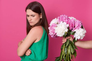 Kaşlarını çatmış yüzü çiçeklerden yüz çeviren çekici esmer genç kadın, katlanmış kolları ile stüdyoda pembe arka plan üzerinde izole ayakta, kenara bakarak, üzgün yüz ifadesi olan.