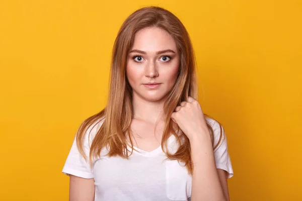 Pretty female with fair hair, dressed casual white t shirt, looking with serious facial expression at camera, being concentrated at something. Studio shot of beautiful woman isolated over studio wall. — Stock Photo, Image